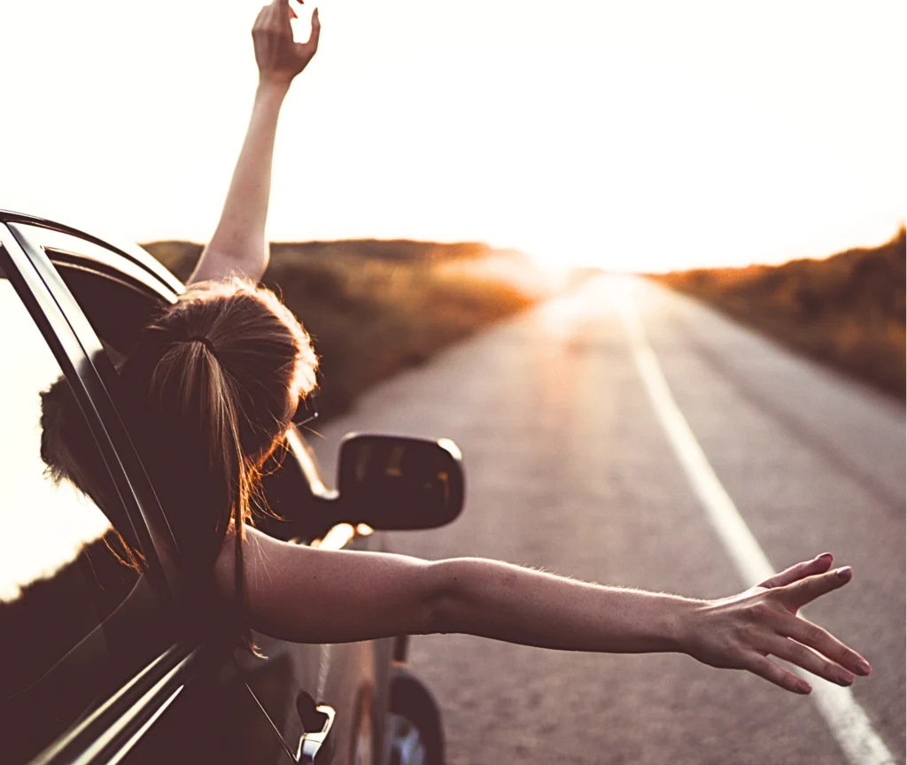 Woman stretching her arms out of a car window at sunset on an open road, representing emotional freedom and personal transformation through coaching.