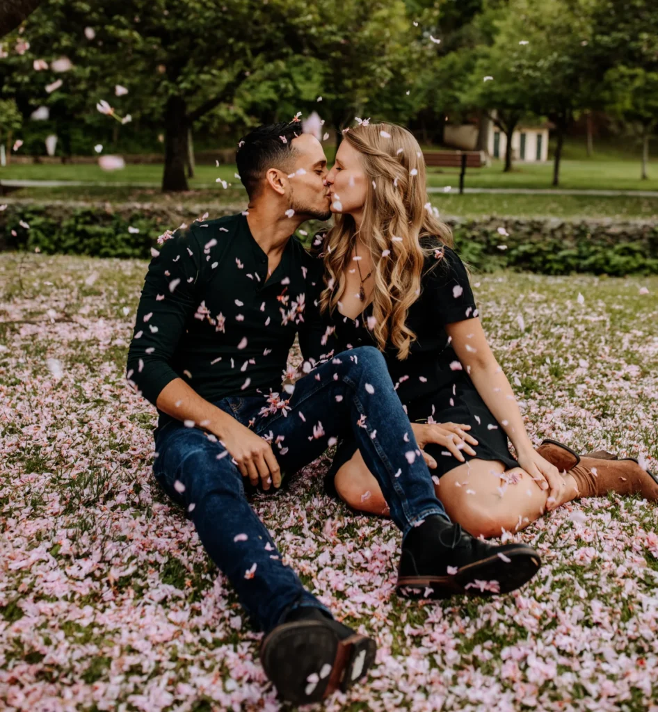 Couple sitting on grass, kissing under falling cherry blossom petals in a park.