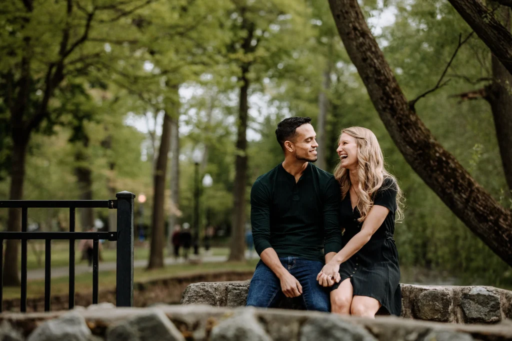 Couple laughing and holding hands on a stone bench in a wooded park.